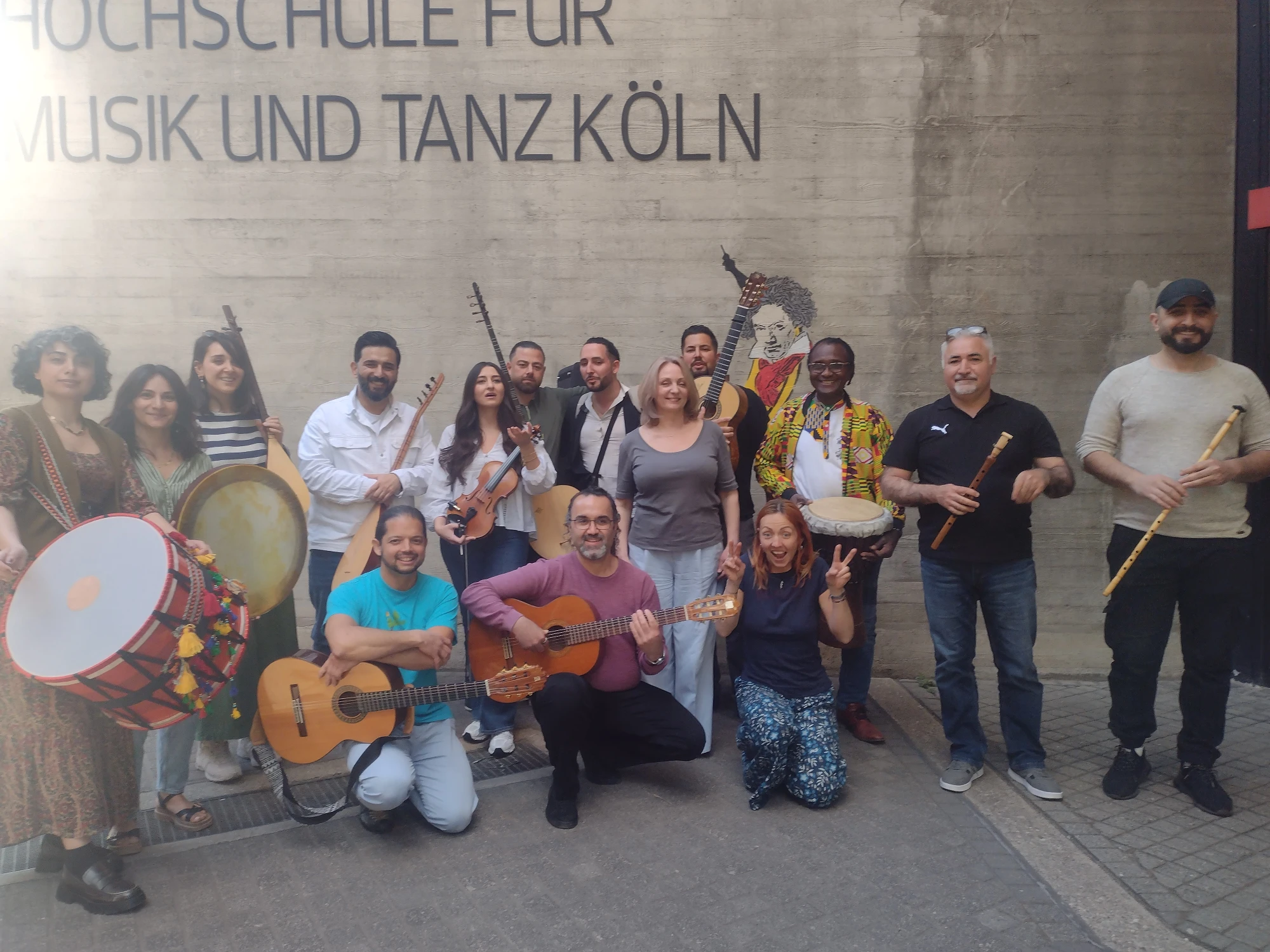 Eine Gruppe von Absolvent:innen des Zertifikatslehrgangs steht mit unterschiedlichsten Instrumenten vor der Hochschule für Musik und Tanz Köln