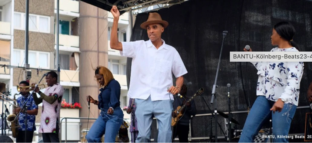 Die Band Bantu auf einer Festival Bühne. Zu sehen sind vier Musikerinnen, vorne steht Ade Bantu der Sänger, gekleidet mit weißem Hemd, hellblauer Hose und einem braunen Hut. Er hält ein Mikro. Daneben stehen eine Sängerin und zwei Bläser.