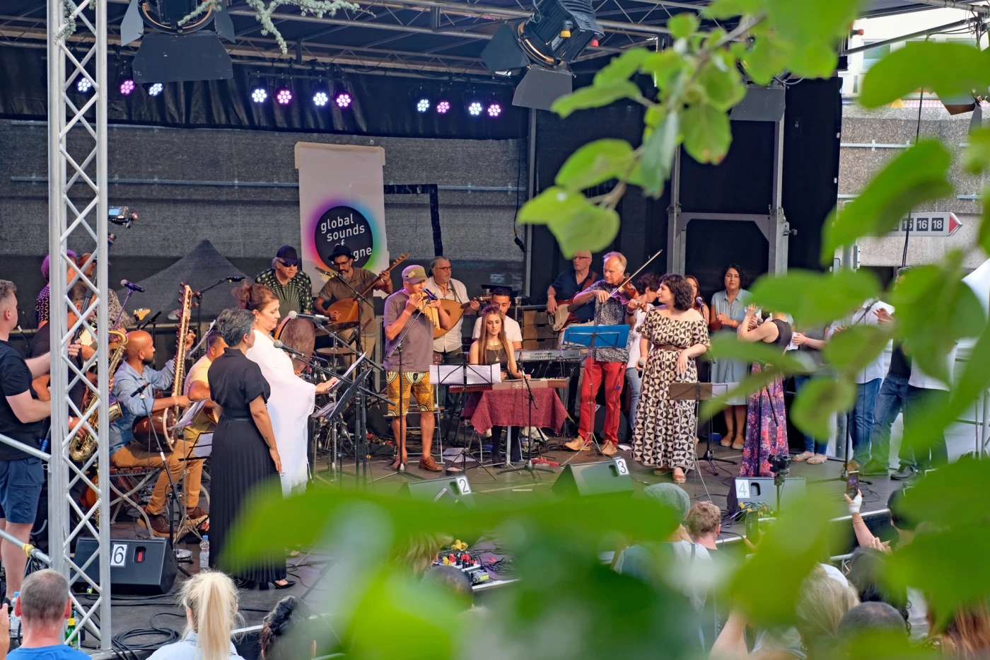 Eine vielfältige Gruppe von Musikern und Sängern tritt auf einer Open-Air-Bühne beim "Global Sounds Cologne"-Event auf. Man sieht verschiedene Instrumente wie Geigen, Gitarren und Flöten.